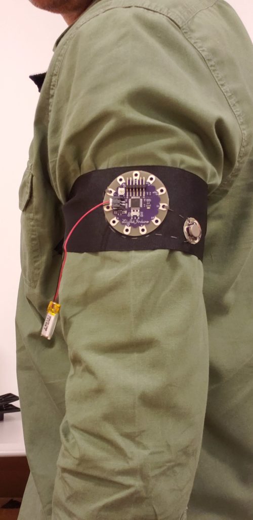 Black ribbon, with Arduino showing, tied around the upper arm of a person wearing an olive green, long-sleeved shirt.