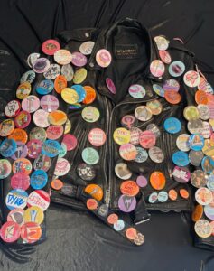 A black leather jacket is laid out on a black background. The jacket is covered in buttons, or badges, with lesbian power slogans, the word dyke, and synonyms for dyke. 