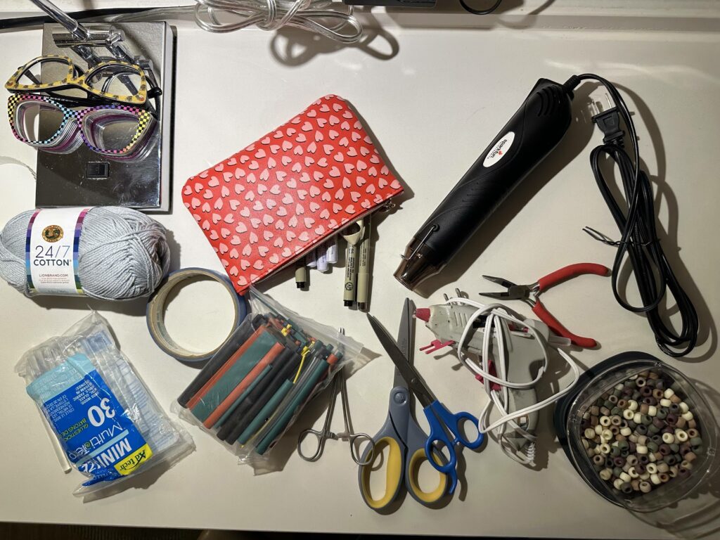 a red pouch with hearts holds paint pens. It sits on a hotel room desk alongside shrink tube, a heat gun, a glue stick, scissors, pliers, and other miscellaneous supplies. 