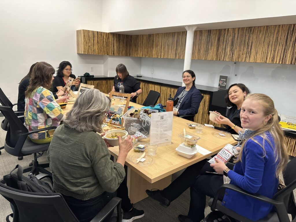 Many women of different ages and races sit around the table smiling, eating, and weaving. 