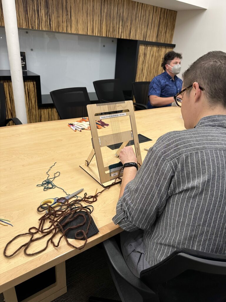 A non binary person with short dark hair and glasses sits with their back to the camera, weaving on th eloom. 